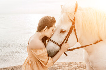 A woman in a dress stands next to a white horse on a beach, with the blue sky and sea in the...