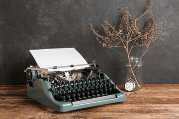 Vintage typewriter, pocket watch and vase with dried flowers on wooden table near black wall