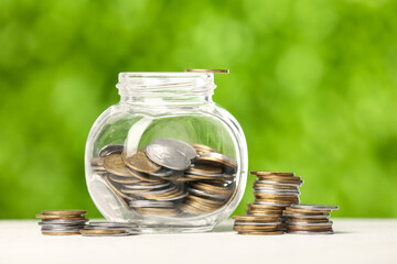 Jar with coins on table against blurred green nature background. Savings concept