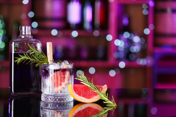 Glass of purple gin and tonic with bottle, grapefruit and rosemary on table in bar against blurred background
