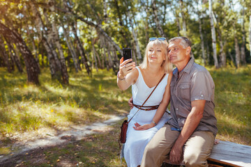 Obraz premium Portrait of senior family couple taking selfie using smartphone in summer forest. Elderly people rest sitting on bench