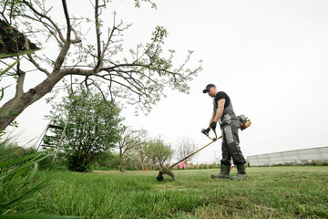 Side view of professional gardener in protective apparel is trimming green grass. Man worker mows weeds with hand electric or petrol lawn trimmer, mower in city park or backyard. Gardening care tools