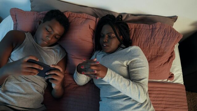 brazilian black child playing on smartphone lying in bed with his mother on mother's day and her tickling him