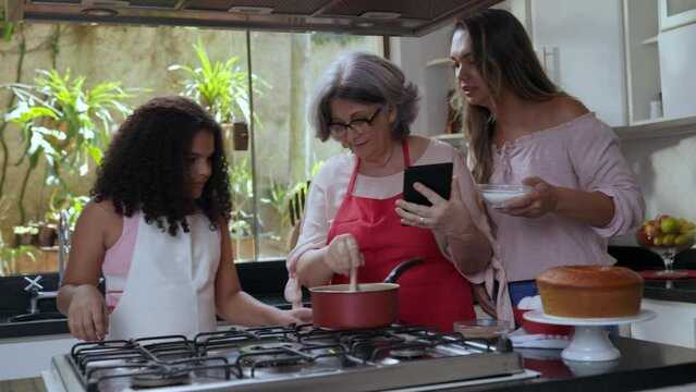 av&oacute; m&atilde;e e filha brasileiras juntas na cozinha preparando um bolo para o dia das m&atilde;es enquanto a av&oacute; confere a receita no celular