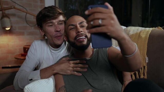 casal gay inter racial latino sentado no sof&aacute; da sala fazendo uma v&iacute;deo chamada no dia dos namorados