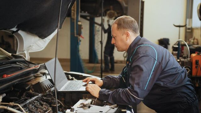 Confident professional car mechanic using a computer laptop to diagnosing and checking up on car engines parts for fixing and repair. Mechanic is repairing an engine in the auto repair