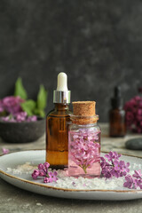 Fototapeta na wymiar Bottles of cosmetic oil with beautiful lilac flowers and sea salt on grey table