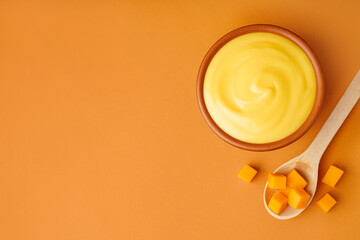 Bowl of tasty cheddar sauce and spoon with cheese cubes on orange background