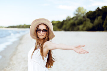 Happy smiling woman in free happiness bliss on ocean beach standing with a hat, sunglasses, and open hands. Portrait of a multicultural female model in white summer dress enjoying nature during travel