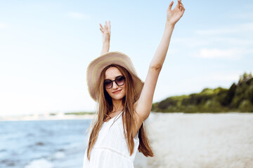 Happy smiling woman in free happiness bliss on ocean beach standing with a hat, sunglasses, and rasing hands. Portrait of a multicultural female model in white summer dress enjoying nature during trav