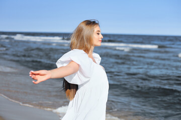 Fototapeta na wymiar Happy, beautiful woman on the ocean beach standing in a white summer dress, open arms.