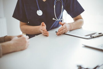 Doctor and patient sitting at the table in clinic while discussing something. The focus is on female physician's hands, close up. Medicine concept.