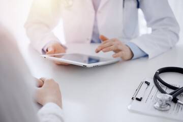 Stethoscope lying on the tablet computer in front of a doctor and patient sitting opposite each other and using tablet computer at the background . Medicine, healthcare concept.