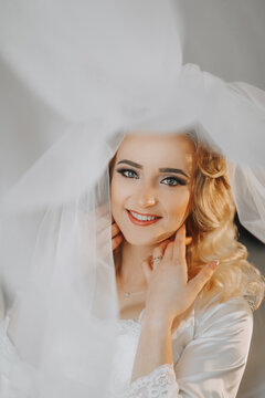 The blonde bride is dressed in a satin robe and lies on a sofa, posing under a veil. Beautiful hair and make-up, open bust. Wedding portrait. Sincere smile