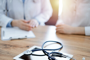 Stethoscope and tablet computer are lying on the wooden table while doctor and patient discussing...