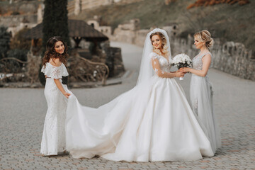 Stylish bridesmaids have fun with the bride in nature and pose to help the bride with the train of...