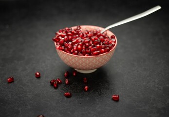 A bowl of red pomegranate  seeds, served with a spoon, and pomegranate seeds around the bowl