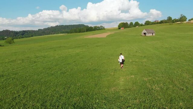 wei&szlig;er Husky Hund und sein Besitzer, ein Junge mit langen Haaren, shorts, einem Achselshirt, einer Umh&auml;ngetasche gehen gemeinsam auf einem Bauernhof Feld auf einer gr&uuml;nen Wiese Gassi im Sommer 4k 