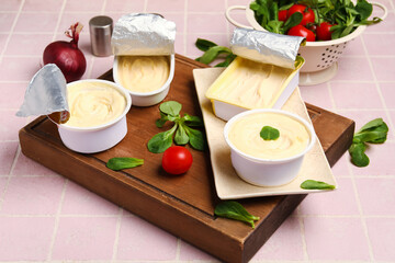 Wooden board with boxes of processed cheese and vegetables on pink tile background