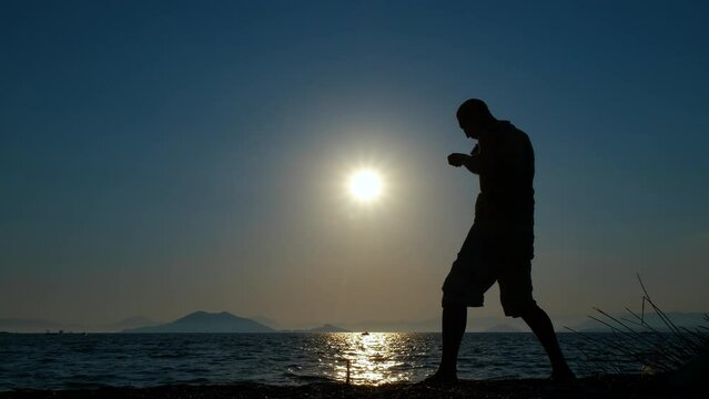 Silhouette man training boxing by ocean. A strong man silhouette training shadow boxing on the empty tropical bay against sunset sky in summer.