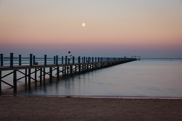Calm sea and wooden dock against the pink sunset sky with a full moon