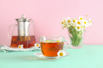 Teapot with cup of natural chamomile tea and flowers on turquoise table near pink wall