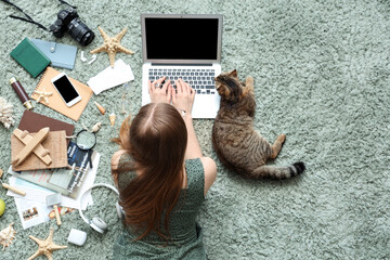 Fototapeta na wymiar Woman with Scottish fold cat and travelling accessories using laptop on green carpet, top view