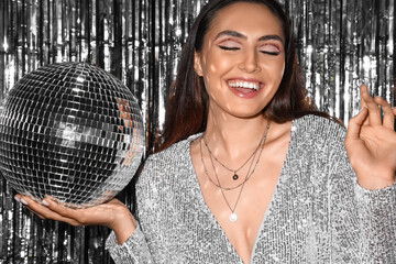 Smiling fashionable woman in sparkling dress with disco ball against silver tinsel