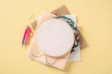 Embroidery concept. Flat lay frame with blank canvas and needlework accessories on pastel yellow...