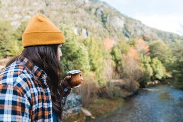 Young latin woman, wearing cozy attire, enjoying yerba mate in nature, contemplating a river and...