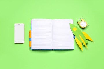 Paper rocket with alarm clock, blank notebook and mobile phone on green background