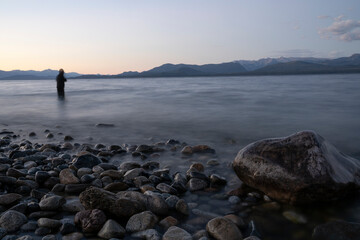Night shot. View the beautiful water blurred effect and blurred fisherman dark silhouette, fishing at nightfall. The rocky shore and mountains in the horizon with a beautiful dusk light.