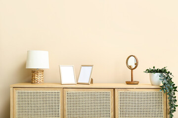 Wooden chest of drawers with lamp, blank pictures, mirror and houseplant near beige wall in room