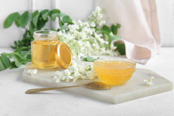 Jar and bowl of honey with flowers of acacia on light background, closeup