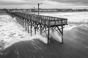 High angle shot of a wooden pier on sea waves crashing against the beach shot in grayscale