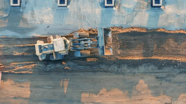 Aerial top down view of backhoe loader and asphalt paver (steam roller or road roller) level the ground and carries out earthworks at the construction site. 