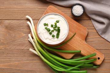 Board and bowl of tasty sour cream with green onion on wooden background