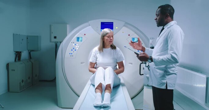 Doctor and patient are having discussion at modern tomography laboratory. African American doctor is holding folder and gesturing. Woman sits at MRI bed prepares for magnetic resonance imaging.
