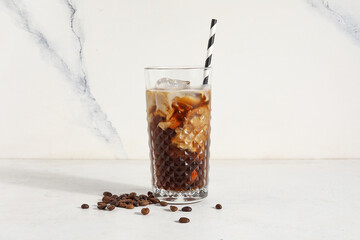 Glass of ice coffee with straw and beans on white table near light grunge wall