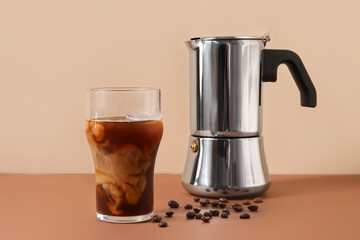 Glass of ice coffee with beans and geyser coffee maker on table near beige wall