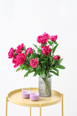 Vase with red peonies and candles on coffee table near white wall