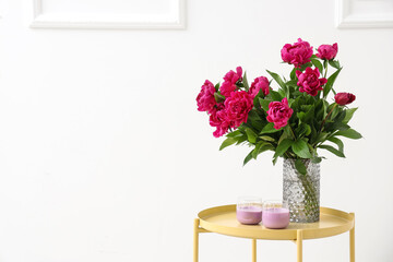 Vase with red peonies and candles on coffee table near white wall