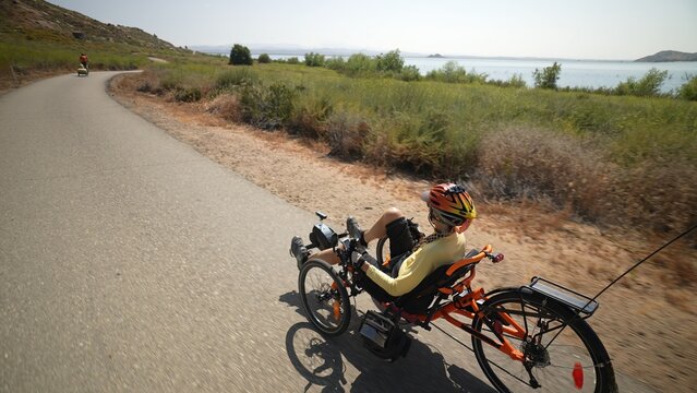 Rear view of elderly senior woman riding recumbent e-bike electric tricycle bicycle beside a lake on a sunny day beside a lake with mountains in the distance.