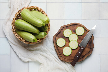 Wicker basket and wooden board with fresh zucchini on white tile background