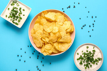 Bowls of tasty sour cream with sliced green onion and potato chips on blue background