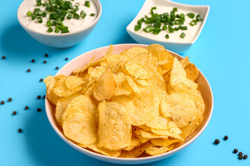 Bowls of tasty sour cream with sliced green onion and potato chips on blue background