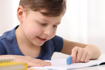 Naklejka na ściany i meble Little boy erasing mistake in his notebook at white desk indoors