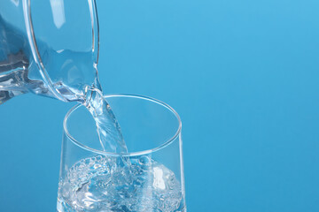 Pouring water from jug into glass on light blue background, closeup. Space for text