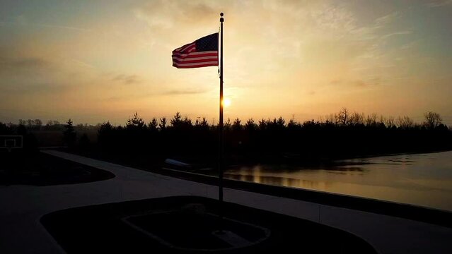 American flag waving on a flagpole against a sunrise, pine trees, and a pond
