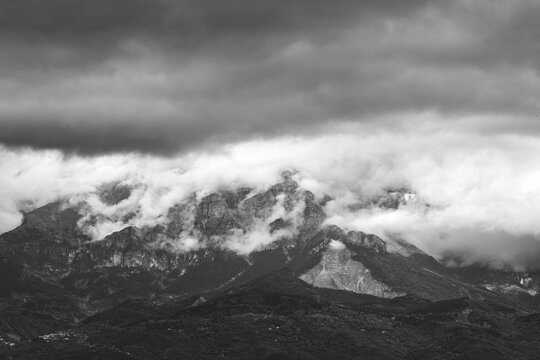 Grayscale of a mountain peak covered in fog with a cloudy dramatic sky in the background © Konstadinos Xenos/Wirestock Creators